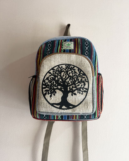 A small woven backpack with a colorful striped border, a beige front panel featuring a black tree of life design, and gray shoulder straps, hanging against a plain off-white wall.